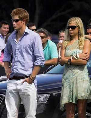 A man and woman stand outdoors, both wearing sunglasses, with a car and crowd in the background.