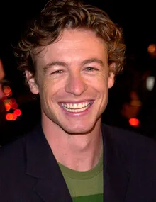 A person with curly hair smiling broadly, wearing a dark jacket and green shirt against a blurred dark background.