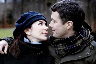 Couple gazing at each other, both wearing winter clothing with dark coats and a plaid scarf, in an outdoor setting.