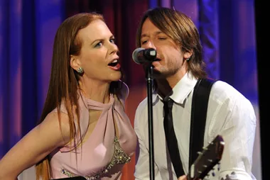 A woman and man perform on stage; she sings energetically into a mic while he plays guitar and sings.