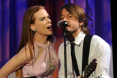 A woman and man perform on stage; she sings energetically into a mic while he plays guitar and sings.