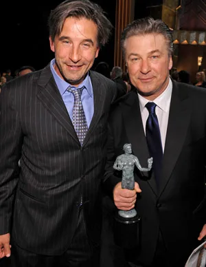 Two men in suits pose together; the man on the right holds an award statue.