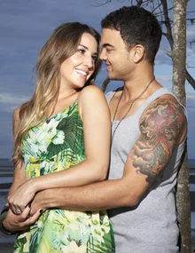 A couple smiling and embracing near a beach, with the man showcasing a colorful arm tattoo.