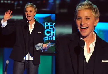 A person with short hair in a suit holding a trophy at an award event, speaking at a microphone and smiling.