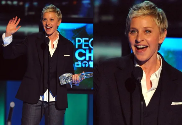 A person with short hair in a suit holding a trophy at an award event, speaking at a microphone and smiling.