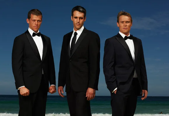 Three men in tuxedos stand on a beach with a blue sky and ocean in the background.