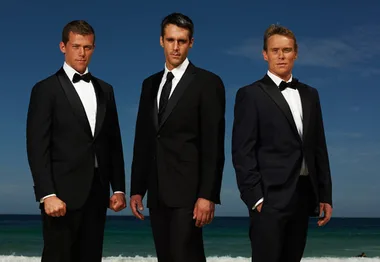 Three men in tuxedos stand on a beach with a blue sky and ocean in the background.