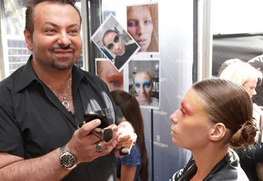 "Makeup artist applying bold eyeshadow to model backstage at fashion week, with makeup looks displayed on the wall."