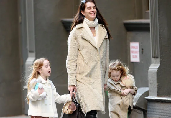 A woman in a light coat walks outside with two young girls, both dressed in warm clothing, carrying bags and dolls.