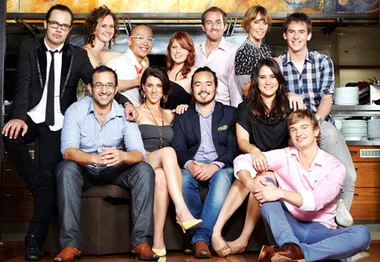 A group of twelve smiling people poses together in a casual indoor setting.