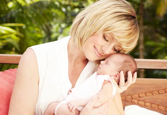 A woman with short blonde hair gently cradles a sleeping baby outdoors, surrounded by lush greenery.