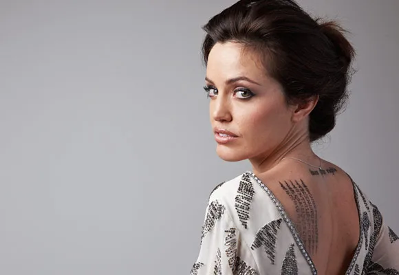 A woman with dark hair and a tattooed back poses in a sequined dress, looking over her shoulder.