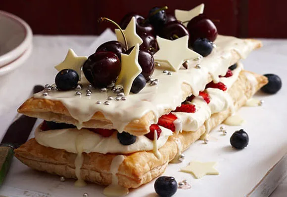 Layered puff pastry dessert with cream, berries, cherries, and white chocolate stars, topped with silver dragees.