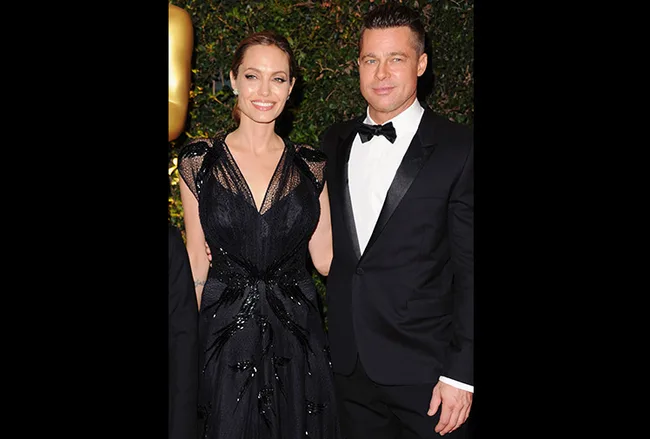 A man and a woman in formal attire smiling at a Hollywood event with greenery in the background.