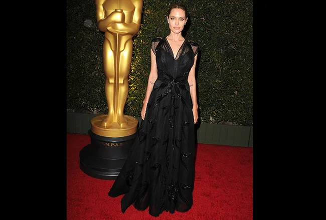 A person in a black dress stands on a red carpet next to a large Oscar statue.