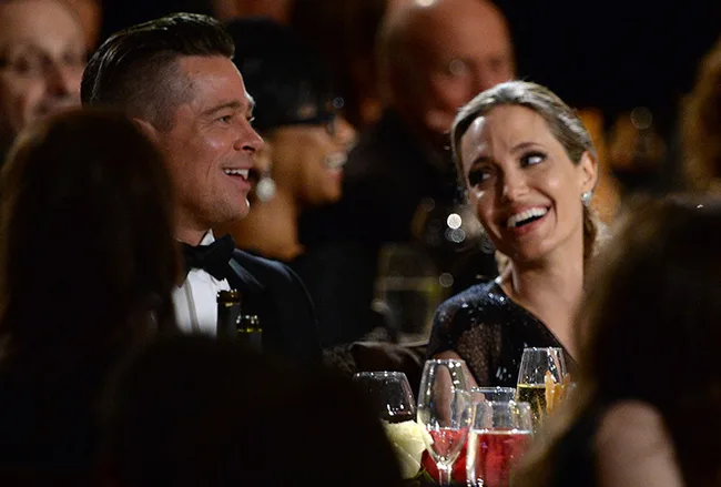 Two well-dressed individuals smiling at a formal event with drinks on the table in front of them.