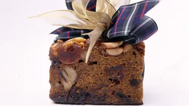 A slice of fruitcake with nuts and dried fruits, wrapped in a decorative tartan ribbon bow on top.