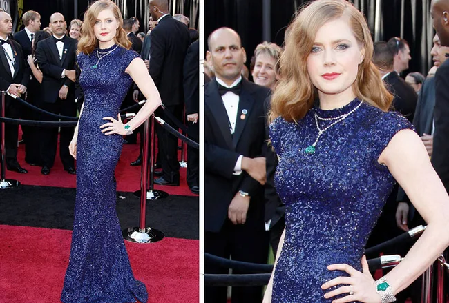 Woman in a sparkling blue gown on a red carpet, posing with one hand on hip, surrounded by onlookers in formal attire.