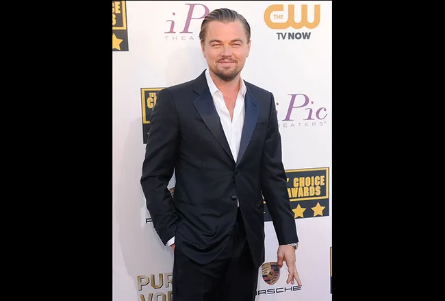 A man in a black suit poses on the red carpet at an award event, with sponsor logos in the background.