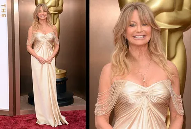 Goldie Hawn at the Oscars red carpet in a flowing gold gown, standing in front of an Oscar statue, smiling.