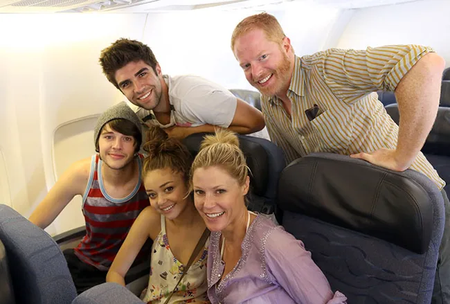 Group of five smiling people sitting in airplane seats, including one man standing behind the seats.