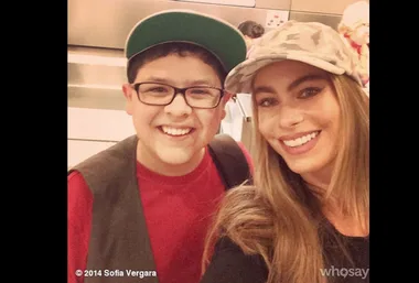 Two smiling individuals taking a selfie, both wearing hats, with blurred background. © 2014 Sofia Vergara.