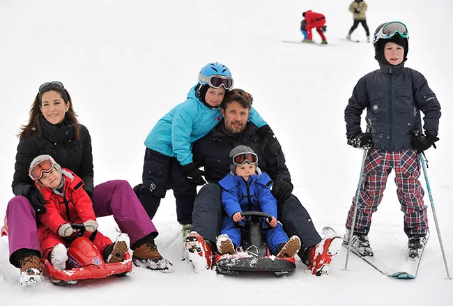 Family enjoying a snowy holiday, dressed in winter gear, with sleds and skiing equipment on a snowy slope.