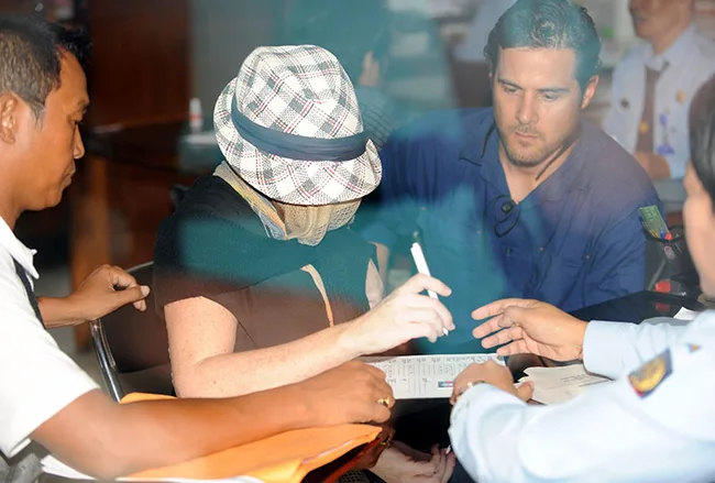 Woman in disguise with a hat and sunglasses, signing documents, surrounded by officials.