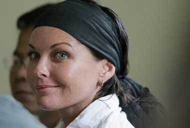 A woman with a black headscarf and white shirt smiles slightly in a close-up portrait, with another person in the background.