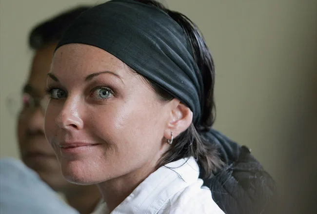 A woman with a black headscarf and white shirt smiles slightly in a close-up portrait, with another person in the background.