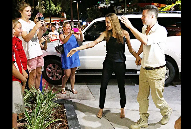 A couple holding hands greets fans taking photos on a city street at night, with a white car in the background.