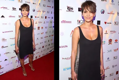 Halle Berry in a black dress at a red carpet event, standing in front of a backdrop with logos.