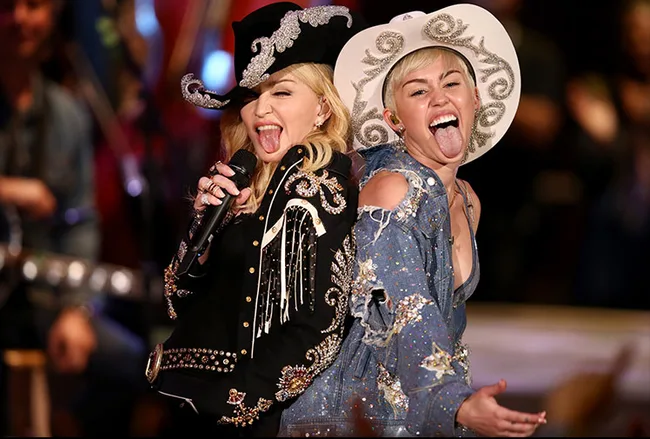 Miley Cyrus and Madonna performing together, wearing embellished cowboy hats and outfits, both sticking out their tongues.