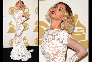 Beyoncé wearing a sheer white lace gown with elegant earrings at a Grammy event, posing confidently against a logo backdrop.