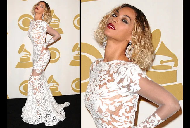 Beyonc&eacute; wearing a sheer white lace gown with elegant earrings at a Grammy event, posing confidently against a logo backdrop.