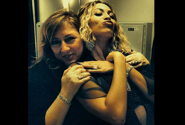 Two women posing closely, both smiling, wearing jewelry. One has curly hair and pouts. Indoor setting.