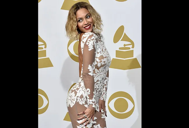 A woman in a white lace dress poses confidently in front of a backdrop with Grammy Award logos.