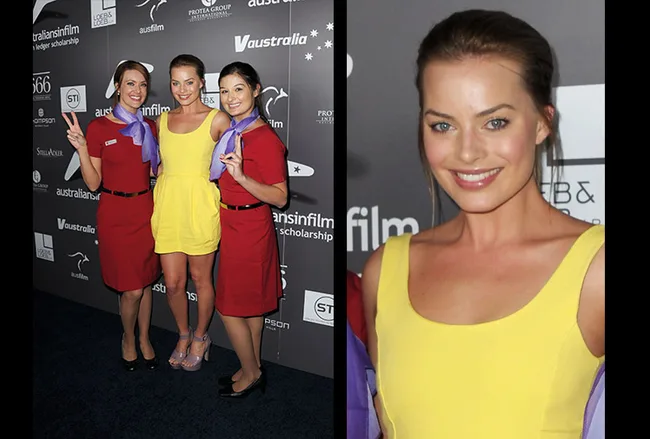 Three women pose together, one in yellow dress, two in red uniforms at an event with sponsor logos on a black backdrop.