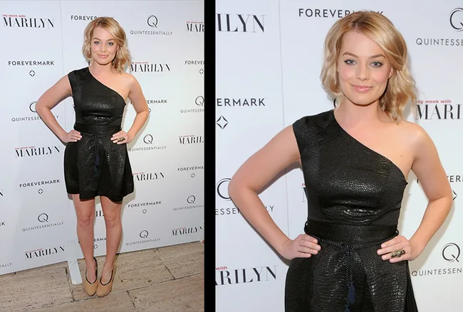 A person in a black one-shoulder dress poses at a red carpet event with a step-and-repeat backdrop behind them.