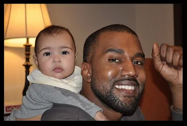Man smiling while holding a baby in a cozy room with a lamp in the background.
