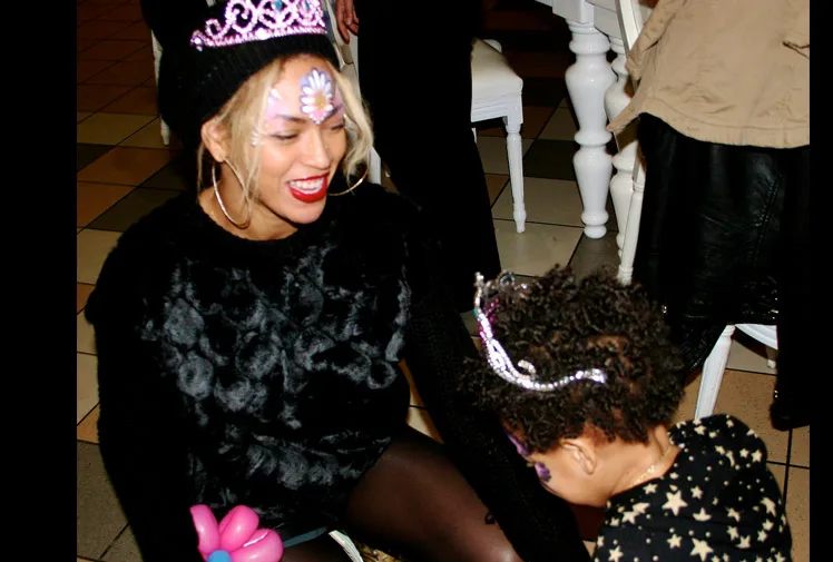 A woman smiling at a child, both wearing tiaras and face paint at a party with festive decor.