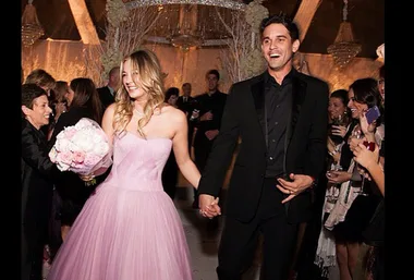 A couple joyfully holding hands at a wedding; the woman in a pink gown, the man in a black suit, surrounded by smiling guests.