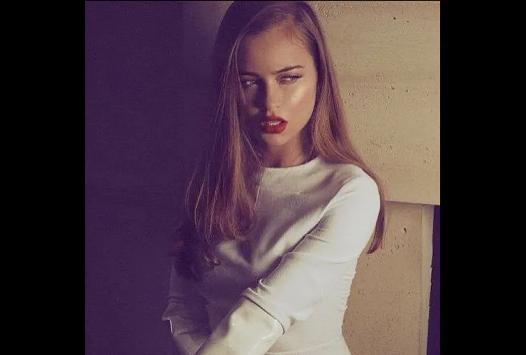 Young woman in a white dress with long hair and red lipstick, posing against a beige wall.
