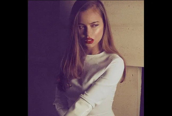 Young woman in a white dress with long hair and red lipstick, posing against a beige wall.