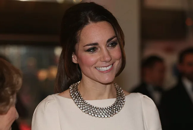 A woman with long hair smiles, wearing a sparkling necklace and a light-colored top, in an elegant indoor setting.