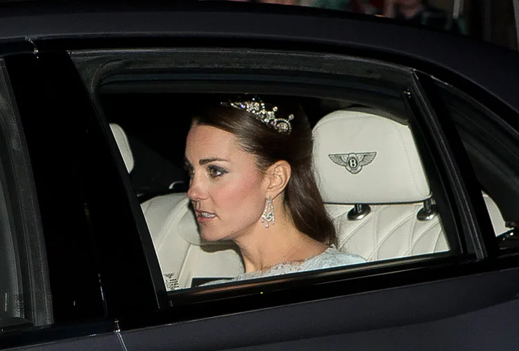 A woman wearing a tiara and earrings sitting in the back seat of a luxury car with a Bentley logo on the headrest.