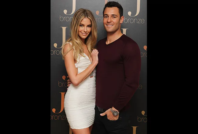 A couple posing together, woman in white dress, man in maroon sweater, in front of a J bronze backdrop.