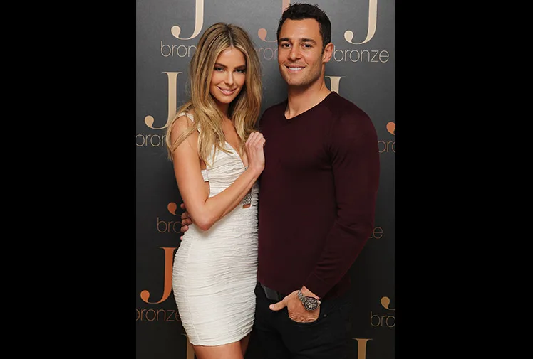 A couple posing together, woman in white dress, man in maroon sweater, in front of a J bronze backdrop.