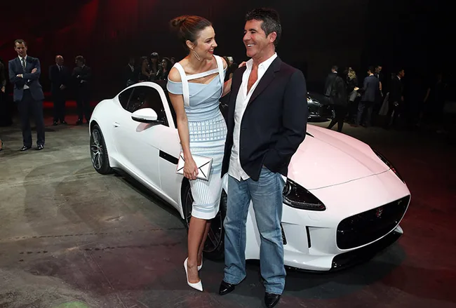 Two people smiling and talking in front of a sleek white sports car at an event.