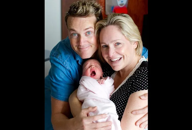 A smiling couple holds a yawning newborn wrapped in a pink blanket.